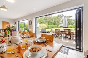Un coin repas avec une table mise pour le petit-déjeuner, une vue sur le jardin grâce à de grandes portes coulissantes et une terrasse aménagée avec du mobilier d’extérieur.