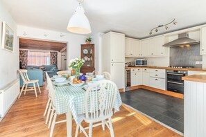 A modern kitchen with white cabinetry, a dining area with a checkered tablecloth, and a cozy living room with a blue sofa. - 3 Bed in Southwold (oc-hsmugg) (Southwold)
