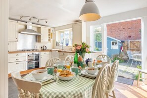 A kitchen with white cabinets, a stove, and a dining table set with breakfast items. - 3 Bed in Southwold (oc-hsmugg) (Southwold)