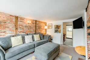 A living room with a grey sofa, a small ottoman, a brick wall, a flat-screen TV, and a dining area in the background. - 3 Bed in Wangford (oc-hchurc) (Wangford)