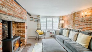 Een woonkamer met een haard van baksteen, een grijze bank, een gele fauteuil en een groot raam met jaloezieën.