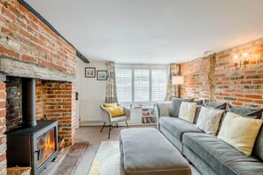 A living room with a brick fireplace, a grey sofa, a yellow chair, and a large window with blinds. - 3 Bed in Wangford (oc-hchurc) (Wangford)