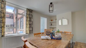 Uma sala de jantar com mesa e cadeiras de madeira, um radiador, janela com cortinas, luminária pendente e uma prateleira com diversos objetos.