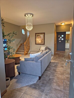 A modern living room with a grey sofa, wooden coffee table, and a chandelier. A staircase leads to another room, and a framed picture hangs on the wall.