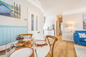A dining area with a table set for two, a blue sofa, and a kitchen in the background. - 2 Bed in Southwold (oc-hgalle) (Southwold)