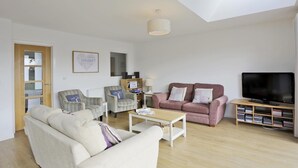 A spacious living room with a high ceiling, a large window, a flat-screen TV, a bookshelf, and a comfortable seating area with a sofa and armchairs. - 3 Bed in Reydon (oc-hspind) (Reydon)