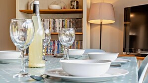 Ein Esstisch mit weißen Tellern, Schüsseln und Weingläsern, eine Flasche Weißwein und eine Lampe. Im Hintergrund steht ein Bücherregal mit Büchern.