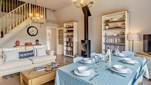 Ein Wohnzimmer mit Sofa, Couchtisch, Esstisch für vier Personen, Bücherregal, Kamin und einer Treppe.