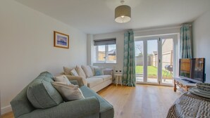 Een woonkamer met een bank, een salontafel en een televisie. Er is een raam met gordijnen en uitzicht op het omliggende landschap.