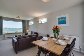 Ein modernes Wohnzimmer mit einer Couch, einem Esstisch für vier Personen und Blick auf das Meer.