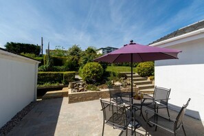 Eine Terrasse mit Tisch und Stühlen, einem violetten Sonnenschirm sowie ein Garten mit Hecke und Vogel-Skulptur.