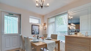 A modern dining area with a wooden table, chairs, and a chandelier. There is a kitchen area with cabinets and a window with blinds. - 1 Bed in Belford (oc-nb677) (Belford)