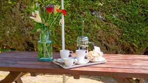 En picnicopsætning på et træbord med en glasvase med blomster, en kaffemaskine, kopper og et fad med bagværk.