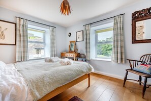 A bedroom with a bed, two windows with curtains, a wooden dresser, a chair, and a mirror on the wall.
