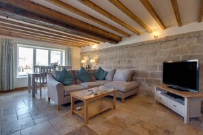 Ein Wohnzimmer mit Sofa, Couchtisch, Fernseher, Steinwand, Holzbalken und einem Essbereich mit Tisch und Stühlen.