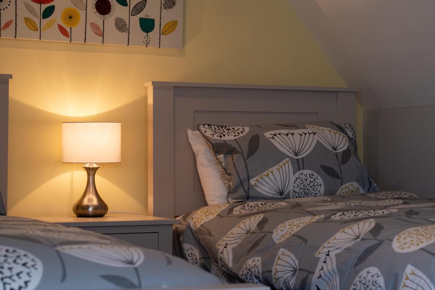 Quarto com cama, mesa de cabeceira, abajur e parede com padrão de folha.