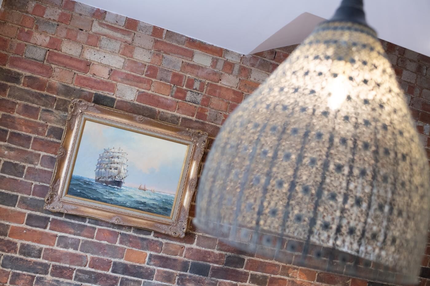 A brick wall with a framed painting of a ship and a textured lamp.