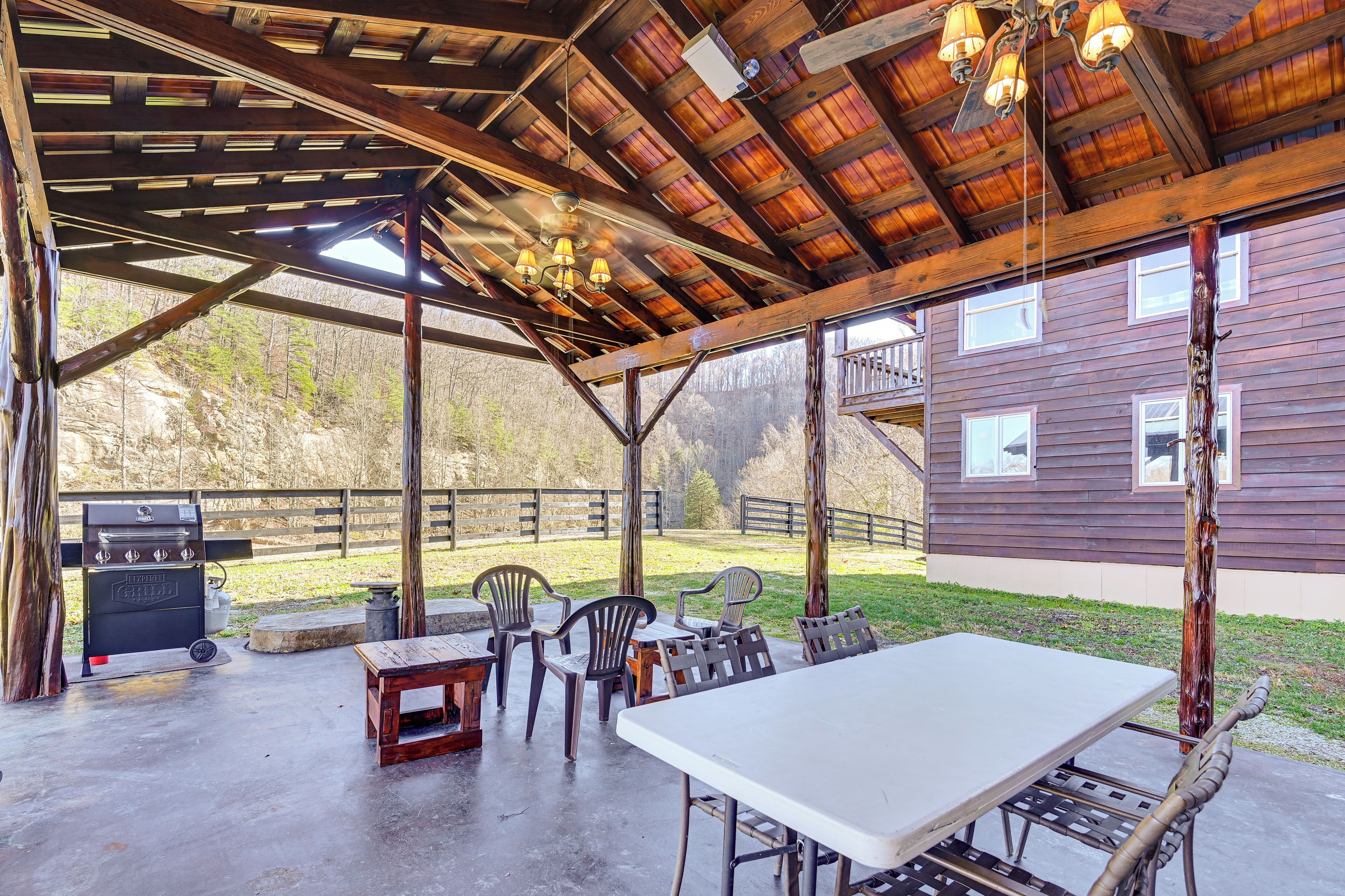 An outdoor covered patio with a table and chairs, a barbecue grill, and a view of a wooden house.