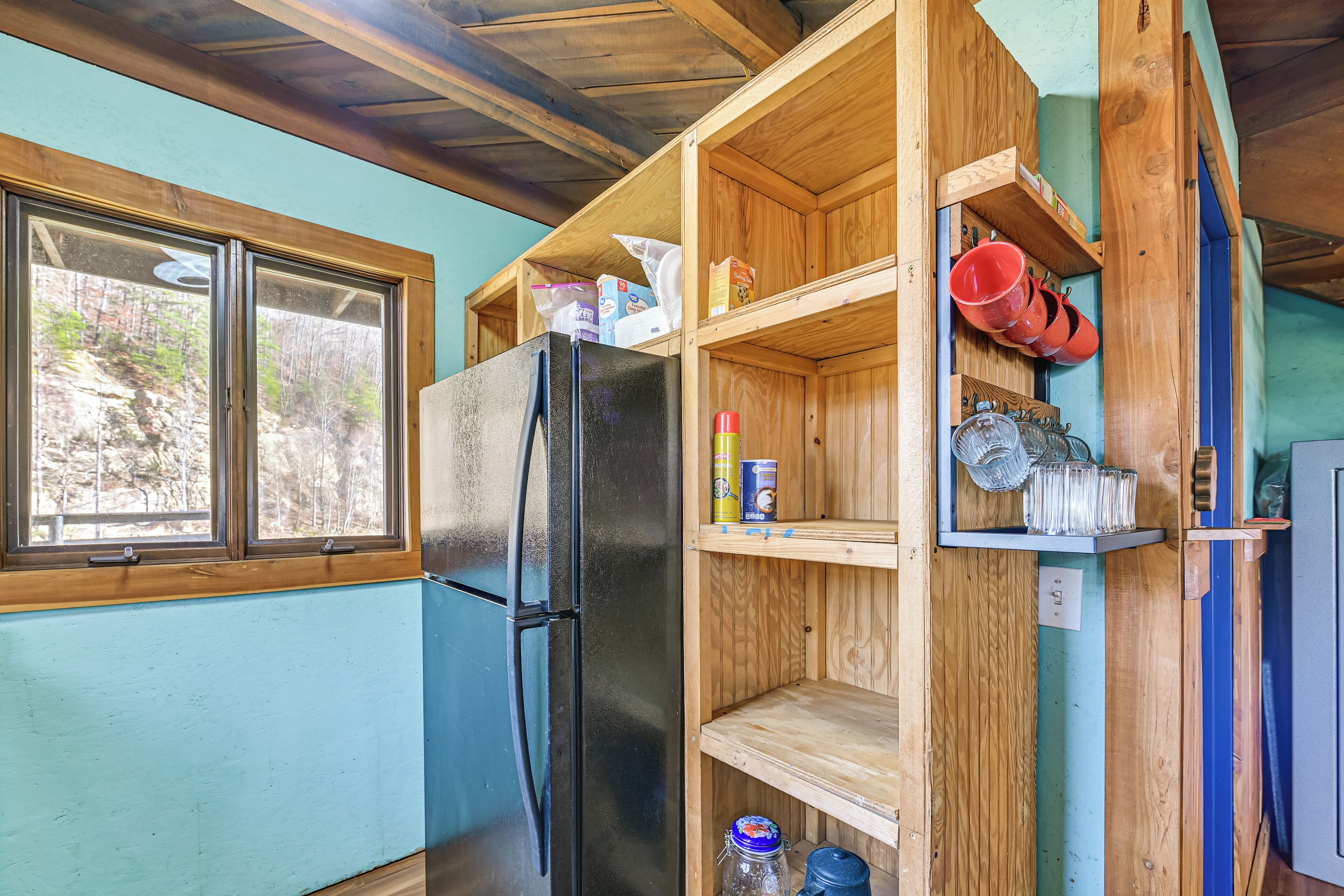 A kitchen with a black refrigerator, wooden shelves, and a window with a view of trees.