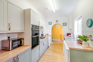 A modern kitchen with white cabinets, a built-in oven, and a microwave. There is a wooden countertop, a sink, and a window with a clock. - 3 Bed in Scarborough (oc-s018) (Scarborough)