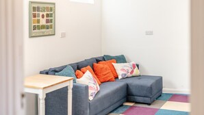 Ein modernes Wohnzimmer mit einem grauen Ecksofa, bunten Kissen, einem kleinen weißen Tisch und einem farbenfrohen Teppich.