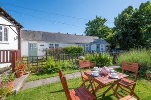 A garden patio with a table set for tea, surrounded by lush greenery and a white building with a green door.