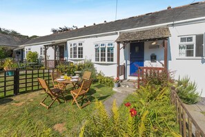 A white cottage with a blue door, a garden with a wooden table and chairs, and a fenced area.