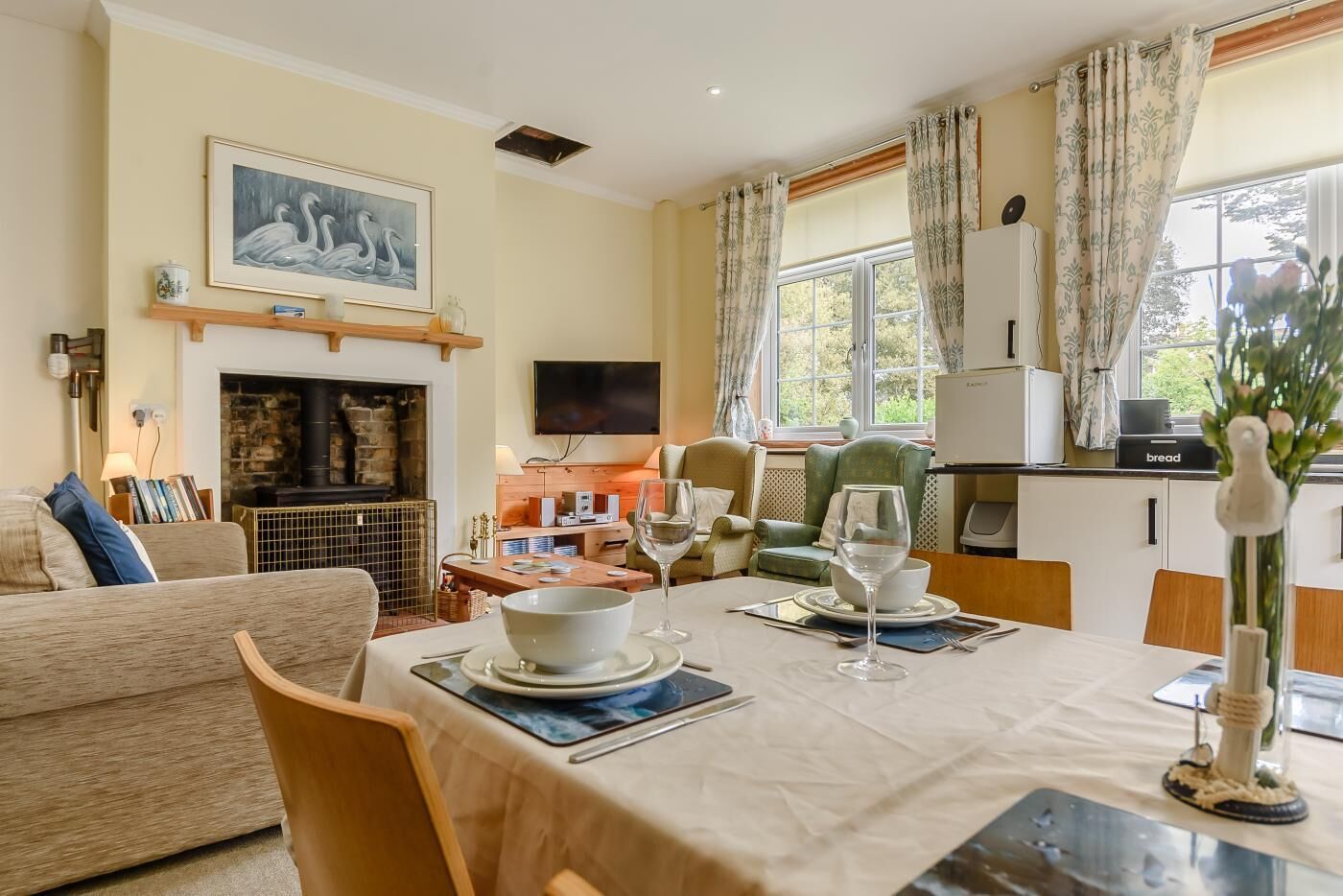 A dining area with a table set for two, a fireplace, a television, and a kitchen area in the background.
