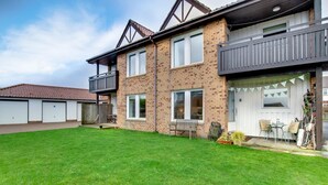 A two-story residential building with a well-maintained lawn, a balcony, and a small patio area with outdoor seating. - 2 Bed in Beadnell (oc-79928) (Beadnell)