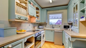 A compact kitchen with light blue cabinets, a wooden countertop, a built-in oven, a dishwasher, and a window with a view of a house. - 2 Bed in Beadnell (oc-79928) (Beadnell)