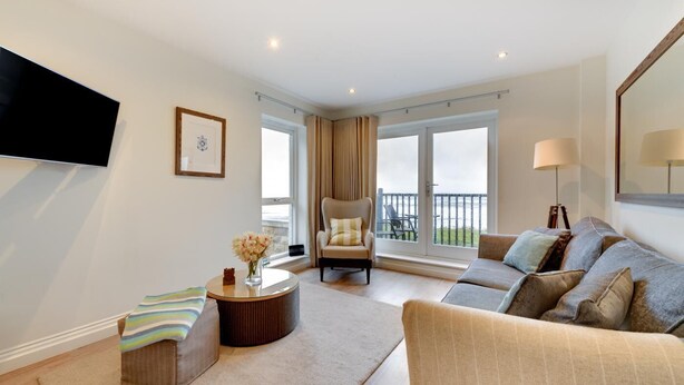 Ein modernes Wohnzimmer mit Sofa, Sessel und rundem Couchtisch. An der Wand ist ein Fernseher angebracht, ein Fenster mit Vorhängen und ein Balkon mit Gartenmöbeln.