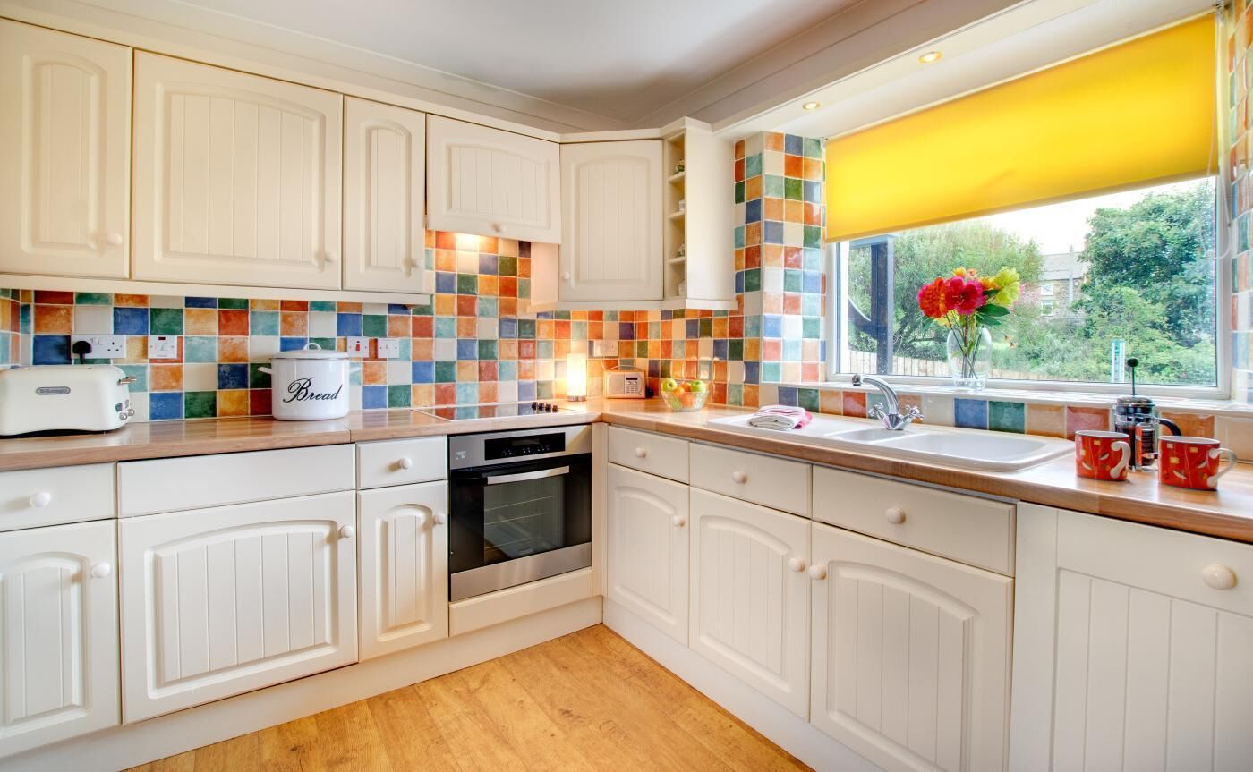 A kitchen with white cabinets, a colorful backsplash, a bread maker, a toaster, and a window with a yellow blind.
