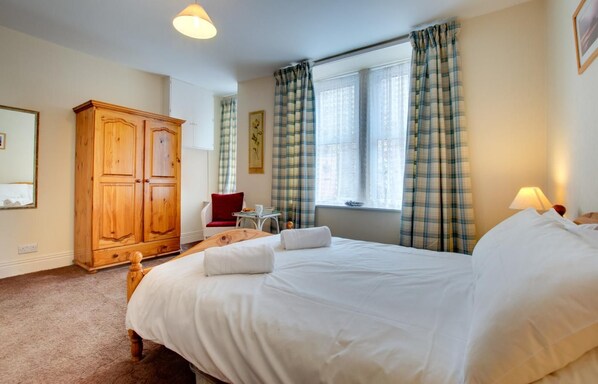 A hotel room with a large bed, a wooden wardrobe, a small table with a chair, and a window with curtains.