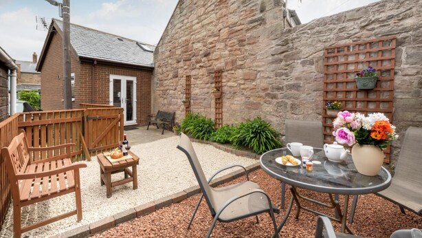 Un espace patio avec une table préparée pour le thé, entouré de murs en pierre et d’une clôture en bois.