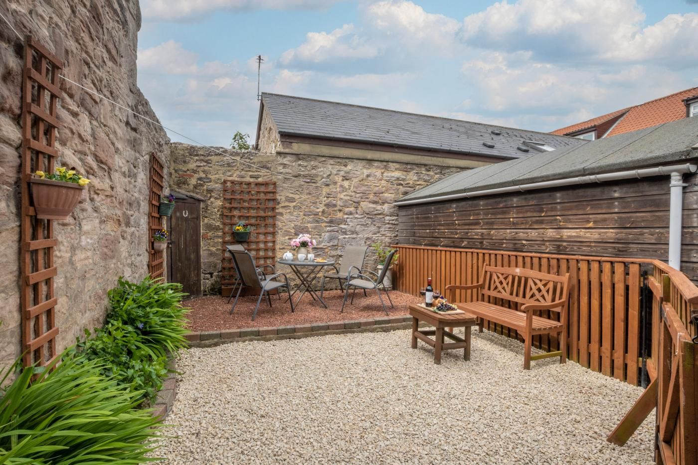 Un bâtiment en pierre avec un toit en pente, une clôture en bois et une terrasse en gravier aménagée avec du mobilier d’extérieur.