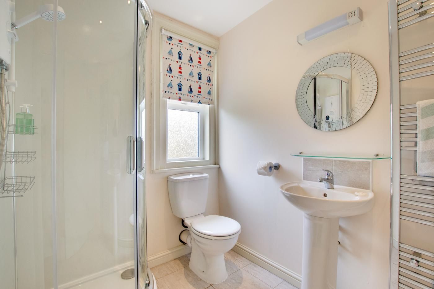Ein Badezimmer mit Duschkabine aus Glas, Toilette, Waschbecken mit Spiegel darüber und einem Fenster mit Segelboot-Motiv.