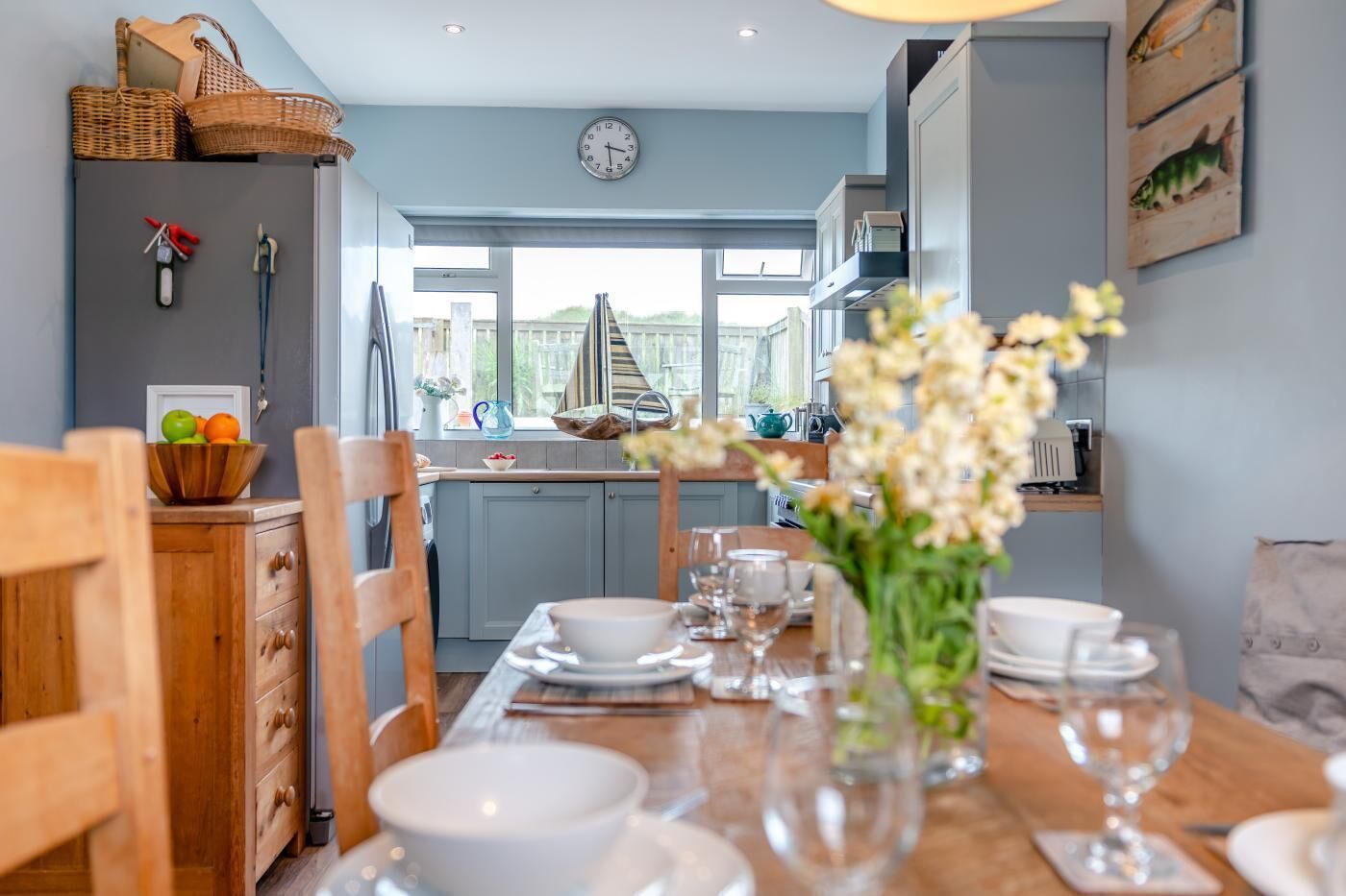 Un coin repas avec une table dressée pour quatre personnes, une cuisine en arrière-plan et une horloge au mur.