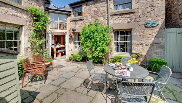 Ein Steinhaus mit einer Terrasse, die Sitzgelegenheiten und einen Tisch mit einem Blumenstrauß bietet.