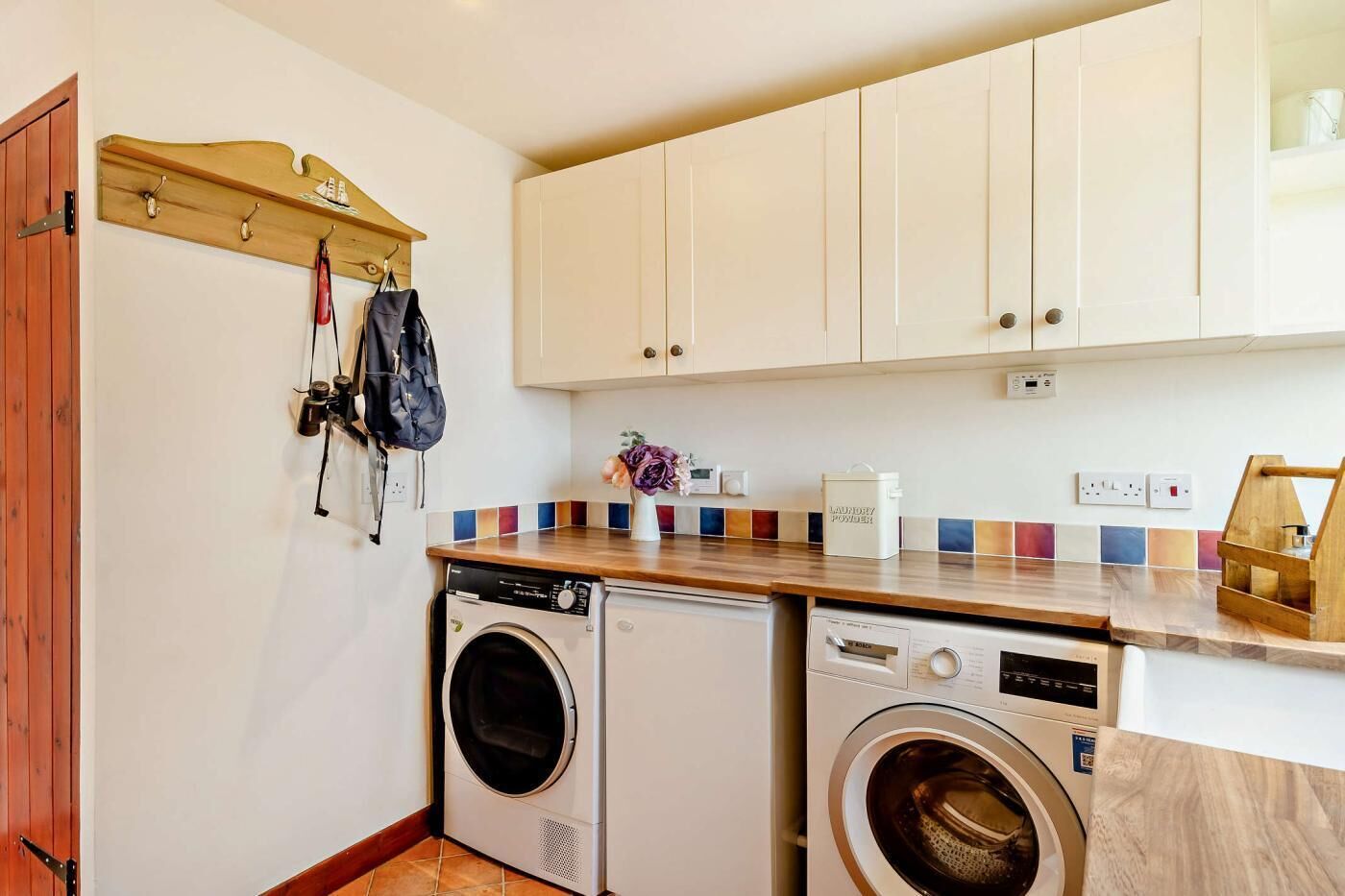 A compact laundry room with two washing machines, a wooden drying rack, and a tiled backsplash.