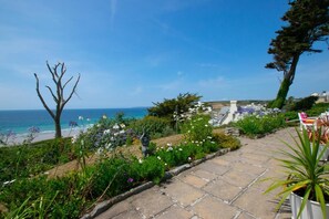 Un jardin avec un sentier en pierre menant à un espace aménagé avec des sièges, d’où l’on a une vue sur la mer.