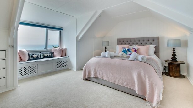Un dormitorio con techo inclinado, cama con cabecera capitonada, asiento junto a la ventana con cojines y una mesita redonda con lámpara.