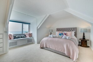A bedroom with a sloped ceiling, a bed with a tufted headboard, a window seat with cushions, and a small round table with a lamp. - 4 Bed in Praa Sands (oc-poohco) (Praa Sands)