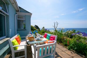 A table set outdoors with a view of the sea, a white bench with colorful cushions, and a house with a blue exterior.