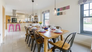 A modern kitchen with a dining area, featuring a wooden table set for a meal, a kitchen island with stools, and a view through a large window. - 4 Bed in Mundesley (oc-1983) (Mundesley)