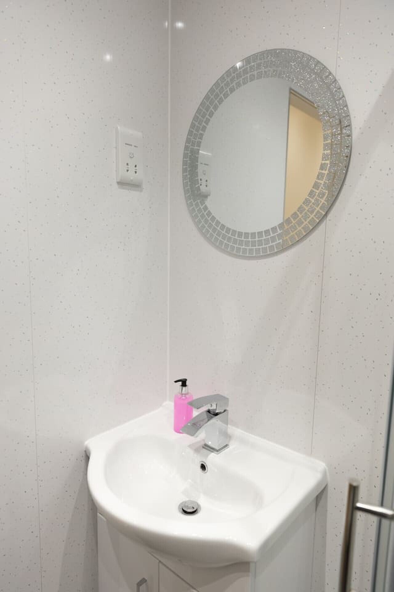 Un baño con un lavamanos blanco, un espejo redondo con un borde de mosaico y un dispensador de jabón rosa.