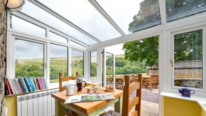 Un invernadero con una mesa dispuesta para el desayuno, libros en una estantería y vistas al campo.