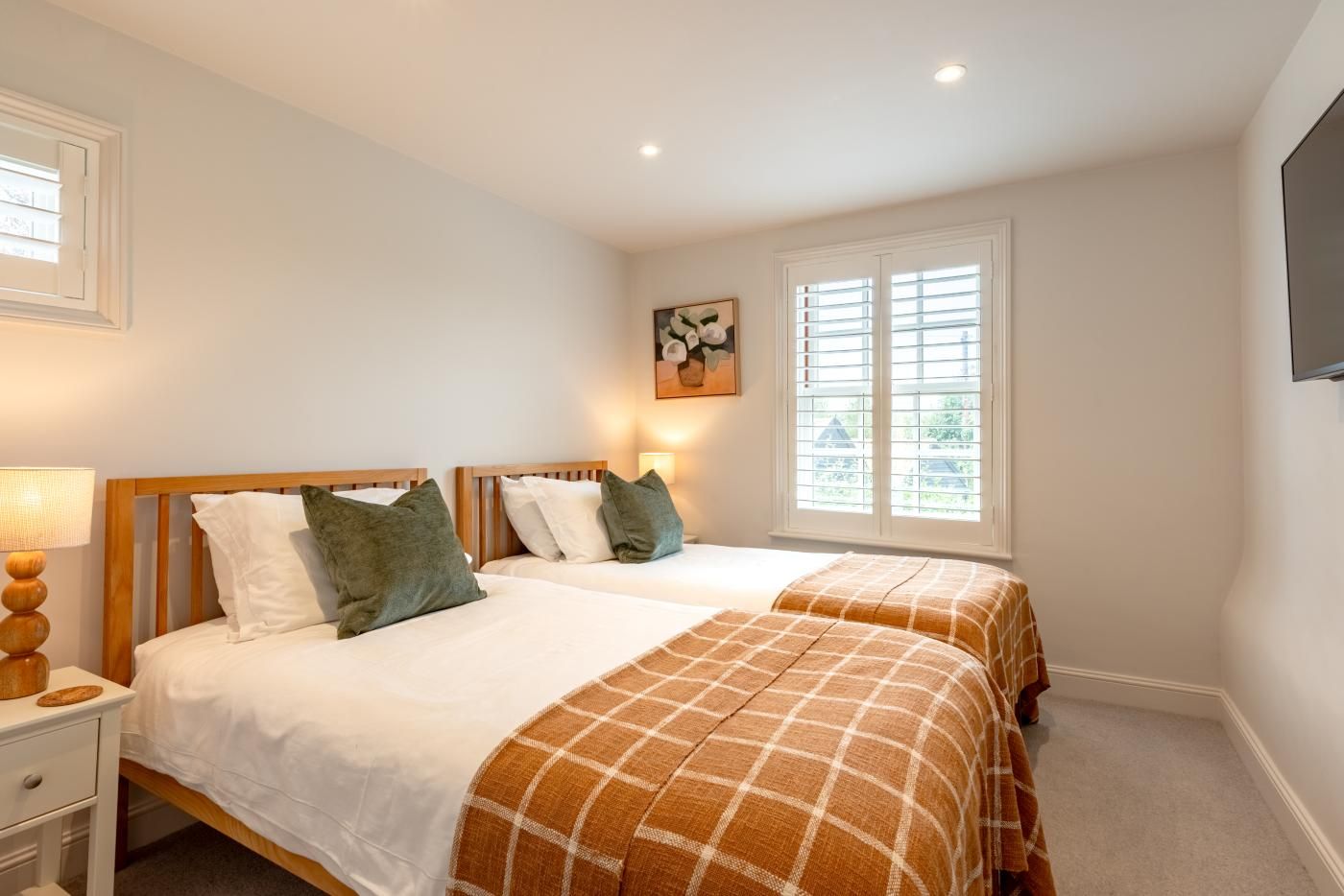 Une chambre d’hôtel avec deux lits, une table de chevet, une lampe, un tableau et une fenêtre avec des stores.