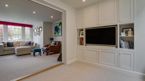 A living room with a sofa, ottoman, and armchair, a flat-screen TV, and built-in shelving with books and decorative items. - 4 Bed in Southwold (oc-hilsi) (Southwold)