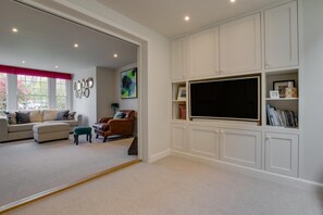 A living room with a sofa, ottoman, and armchair, a flat-screen TV, and built-in shelving with books and decorative items. - 4 Bed in Southwold (oc-hilsi) (Southwold)