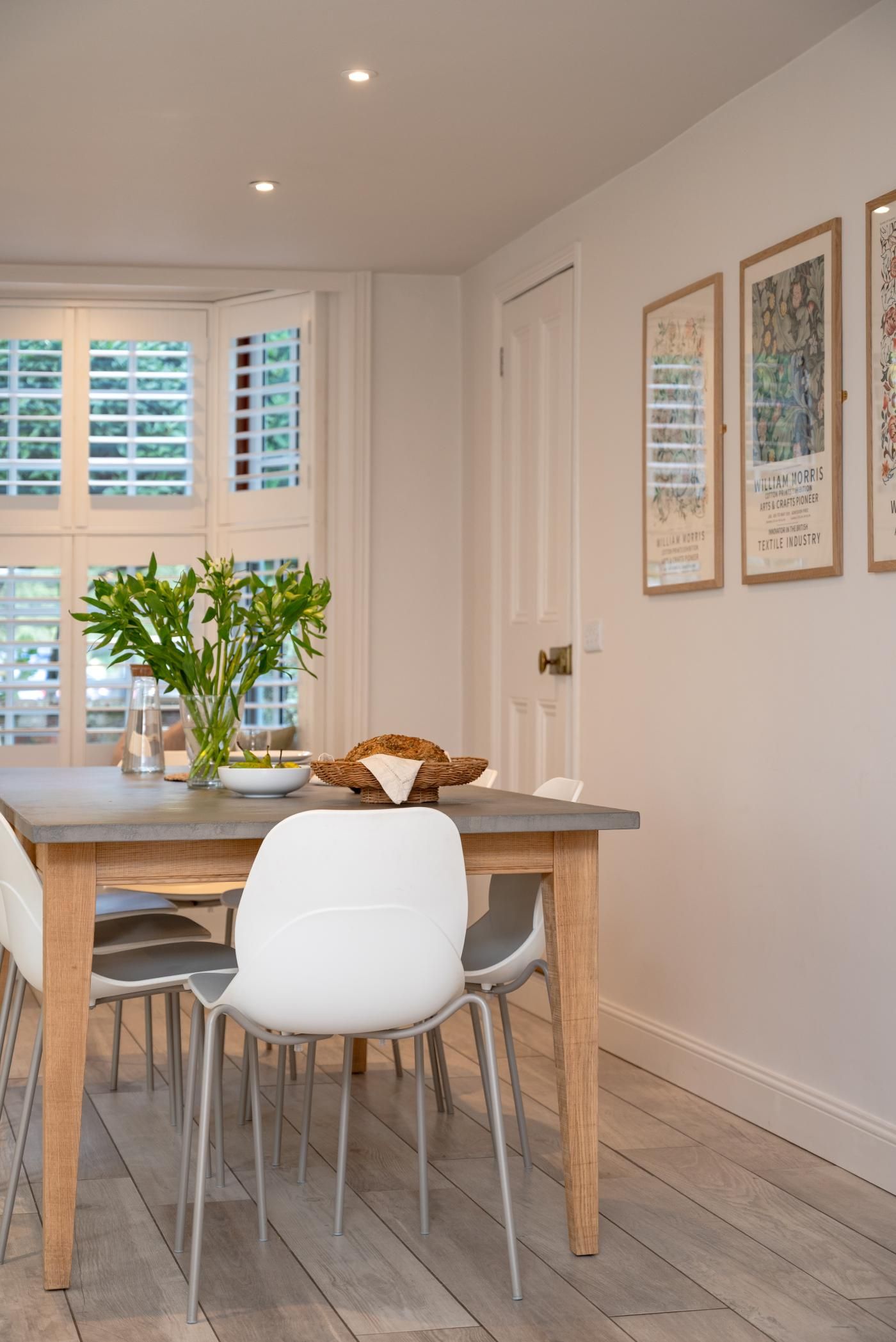 A dining area with a wooden table, white chairs, and a vase with green plants.